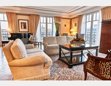 Luxuriöses Wohnzimmer mit beigen Sofas und Vorhängen vor großen Fensterfronten, die auf eine Terrasse mit Blick auf das Brandenburger Tor führen. Ein schwarzer Flügel, ein dunkel lackierter Couchtisch mit Obstschale und Teppich bestimmen die stilvolle Einrichtung in einem klassisch-eleganten Ambiente.