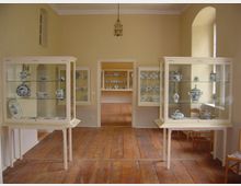 Raum in einem Museum mit gelb gestrichenen Wänden, Holzboden und mehreren Glasvitrinen, die blau-weißes Porzellan wie Teller, Tassen und Vasen ausstellen. Im Hintergrund ist ein weiterer Raum mit zusätzlichen Porzellanexponaten sichtbar.