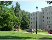 Ein Wohnhaus mit vier Etagen und heller Fassade steht am Rand einer grünen Parkanlage mit Rasen und Bäumen. Im Vordergrund verläuft eine Straße mit geparkten Autos, und eine Straßenlaterne ist sichtbar.