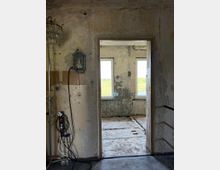 Blick durch eine Türöffnung in einen renovierungsbedürftigen Raum mit bröckelnden Wänden und einem rohen Betonboden. Im Vordergrund linken ist eine Wandlampe, Stromkabel hängen an der Wand, während durch zwei Fenster im Hintergrund grüne Landschaft sichtbar ist.