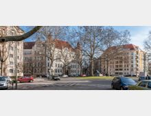 Straßenszene mit Wohn- und Bürogebäuden im neoklassizistischen Stil, darunter ein großes Gebäude mit heller Fassade und rotem Dach. Kahle Bäume und parkende Autos säumen die Straße, während ein kleiner Kreisverkehr mit Grünfläche in der Mitte sichtbar ist.