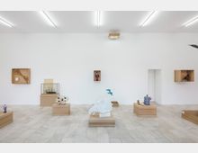 Ein minimalistischer Ausstellungsraum mit hellen Wänden und gefliesten Böden, in dem verschiedene Objekte auf braunen Sockeln und in Wandnischen präsentiert werden. Zu den Exponaten gehören eine weiße Skulptur, bunte Figuren und dekorative Gegenstände, die kunstvoll arrangiert sind.