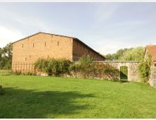 Ein großes Gebäude aus roten Ziegelsteinen steht auf einer grünen Wiese. Davor befindet sich eine Steinmauer mit einem Rundbogendurchgang, die teilweise von Pflanzen und roten Blumen überwachsen ist.
