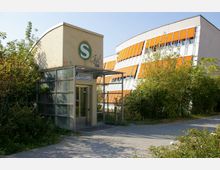 Eingangsbereich einer S-Bahn-Station mit gläsernem Vorbau und grünem „S“-Logo auf einem cremefarbenen Gebäude. Daneben ist ein modernes Gebäude mit orangefarbenen Fensterläden zu sehen, umgeben von Gehwegen und viel Grün.