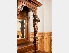 Detailansicht eines reich verzierten Holzinterieurs mit großem Rahmen, Spiegel und geschnitzter Figur als Stütze. Im Hintergrund sind helle Wände und holzvertäfelte Wandpaneele zu sehen.