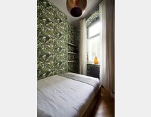 Kleines Schlafzimmer mit tropisch gemusterter Tapete, einem schlichten Bett mit weißer Bettwäsche, schmalen Wandregalen, einem Fenster mit weißen Vorhängen und einer orangefarbenen Vase auf der Fensterbank.