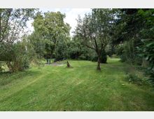 Grüne Gartenlandschaft mit kurz gemähter Wiese, umgeben von Bäumen und Sträuchern. Im Hintergrund ist eine kleine Sitzgruppe mit Stühlen und einem Tisch zu sehen, die auf einer gepflasterten Fläche stehen.