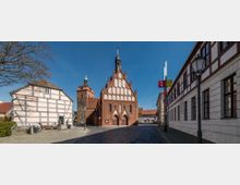Straßenszene mit einem gotischen Backsteingebäude mit spitzbogigen Fenstern und Giebelelementen in der Mitte, flankiert von einem Fachwerkhaus links und einem historischen Gebäude rechts. Im Hintergrund ist ein Kirchturm mit grünlicher Spitze zu sehen, der Himmel ist klar und blau.