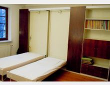 Ein schlichtes Schlafzimmer mit zwei Einzelbetten vor einem großen, wandfüllenden Einbauschrank in dunklem Holz, teils mit offenen Regalen und Schubladen. Links befindet sich ein Fenster mit Blick nach draußen und darunter ein Heizkörper; der Boden ist aus Holz.