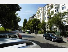 Straßenszene mit mehrstöckigen hellen Wohngebäuden auf der rechten Seite, Balkonen mit Pflanzen und einer Baumreihe entlang des Gehwegs. Links parken mehrere Autos auf dem Kopfsteinpflaster, und im Hintergrund sieht man weitere Gebäude und Bäume unter einem klaren, blauen Himmel.