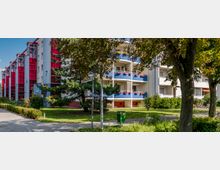 Mehrstöckige Wohngebäude mit bunten Balkonen, blauen und roten Akzenten, umgeben von Bäumen und gepflegten Grünflächen. Gehwege und ein Mülleimer sind im Vordergrund sichtbar, im Hintergrund befinden sich nummerierte Eingänge mit roten Glasfassaden.