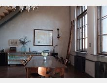 Wohnzimmer oder Essbereich in einem industriellen Stil mit hohen Fenstern, Holztisch und Stühlen. An der Wand befinden sich ein Regal mit Gläsern und Büchern, eine Leiter, Wandlampen und Bilderrahmen.