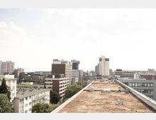 Blick auf eine städtische Skyline mit mehreren Wohn- und Bürogebäuden, darunter Hochhäuser und niedrige Gebäude. Im Vordergrund ist ein begrüntes Flachdach eines Gebäudes zu sehen, umgeben von Bäumen und weiteren Gebäuden.