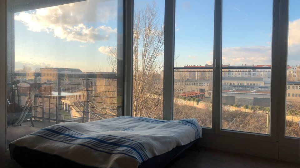 Ein minimalistisches Schlafzimmer mit einem niedrigen Bett vor einer großen Fensterfront, die einen urbanen Ausblick auf Gebäude, Bäume und einen blauen Himmel mit Wolken bietet. Außerhalb der Fenster ist ein Balkon mit einem Geländer sichtbar.