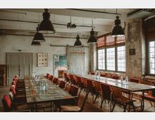 Industriell gestalteter Besprechungsraum mit großen, rustikalen Holztischen, roten Polsterstühlen und schwarzen Hängelampen. Der Raum hat hohe Decken, unverputzte Wände, große Fenster mit Jalousien und eine Schiebetür aus Holz.