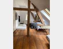 Ein einfacher Dachbodenraum mit Holzbalken und Holzfußboden, ausgestattet mit mehreren Betten, einfachen weißen Laken und einem großen Dachfenster, das Tageslicht hereinlässt. Kleidungsstücke hängen an der Wand, und ein kleines Fenster bietet Blick ins Freie.