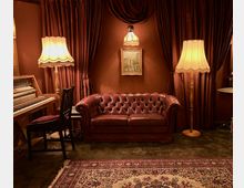 Ein gemütlicher Raum mit dunkelroten Vorhängen, einer roten Ledercouch im Chesterfield-Stil, zwei Stehlampen mit Fransen, einem antiken Teppich und einem gemalten Bild an der Wand. Links steht ein Klavier mit einem schwarzen Stuhl.