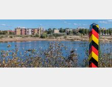 Blick auf einen Fluss mit einem schwarz-rot-gelben Grenzpfosten im Vordergrund, dahinter eine Uferlandschaft mit Bäumen und mehrfarbigen Wohngebäuden im Hintergrund. Im Fluss ist ein kleines Boot mit zwei Personen zu sehen.