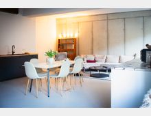 Offener Wohn- und Küchenbereich im modernen Industriestil mit Betonwand, schwarzer Küchenzeile und heller Kücheninsel. Davor stehen ein Esstisch mit vier Stühlen sowie eine große helle Eckcouch mit rundem Couchtisch und Regal im Hintergrund.