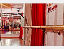 Tanzstudio mit Spiegelwand und roten Vorhängen, einer Ballettstange an der rechten Wand und Holzdetails an der Decke. Der Raum ist mit Holzboden ausgestattet und sanft beleuchtet.