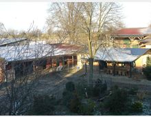 Hofanlage mit mehreren einstöckigen Gebäuden, teils mit roten Ziegeldächern, umgeben von Bäumen und Sträuchern. Im Vordergrund ist ein kleiner Gartenbereich erkennbar, im Hintergrund offene ländliche Umgebung.