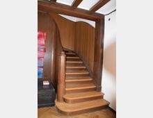 Eine hölzerne Wendeltreppe mit abgerundeten Stufen, eingefasst von einer geschwungenen Holzverkleidung und einem massiven Holzgeländer, steht in einem Raum mit Holzboden und Holzbalkendecke. Links befindet sich ein kleiner, dunkler Beistelltisch mit Büchern darauf.