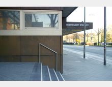 Außenansicht des Bezirksamts Steglitz. Sichtbar sind eine braune Gebäudefassade mit Fenstern, eine Treppe mit Metallgeländer sowie ein Gehweg an einer angrenzenden Straße mit Fahnenmasten und Bäumen im Hintergrund.