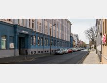 Straßenansicht mit einem blauen Gebäude auf der linken Seite, das durch ein Schild als "Jobcenter" gekennzeichnet ist. Entlang der Straße sind Autos parallel geparkt, rechts ist ein Gehweg mit Pflastersteinen und vereinzelten Bäumen sichtbar.