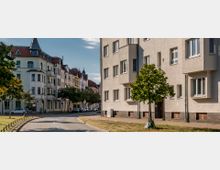 Straßenszene in einem Wohngebiet mit historischen und modernen Wohnhäusern; links stehen mehrstöckige Gebäude mit verzierten Fassaden und roten Dächern, rechts ein Gebäude mit schlichter Putzfassade. In der Mitte verläuft eine gebogene Straße, flankiert von kleinen Bäumen und Gehwegen.