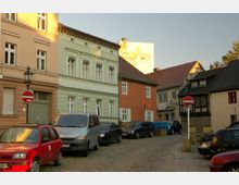 Straßenszene in einer Altstadt mit gepflasterter Fahrbahn, flankiert von historischen Wohnhäusern in verschiedenen Farben, darunter pastellgrün, orange und Fachwerk. Am Straßenrand parken mehrere Autos, und Schilder für ein Einfahrverbot sind sichtbar.