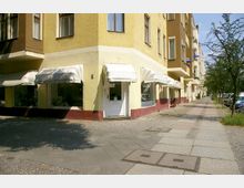 Ecke eines Wohn- und Geschäftsgebäudes mit hellgelber Fassade und roten Sockeln, großen Schaufenstern und weißen Markisen; daneben ein breiter Gehweg mit Pflastersteinen, kleinen Bäumen und parkenden Autos am Straßenrand.