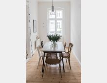 Esszimmer in einem minimalistischen Stil mit einem rechteckigen Tisch aus hellem Material, umgeben von vier unterschiedlichen Stühlen. Im Hintergrund befindet sich ein großes Fenster mit weißen Sprossen, und auf dem Tisch steht eine Vase mit frischen grünen Pflanzen.