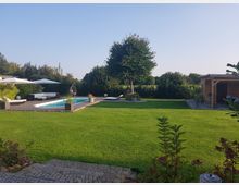Großer Garten mit gepflegtem Rasen, einem rechteckigen Pool umgeben von einer Terrasse mit Sonnenliegen und Sonnenschirmen. Im Hintergrund stehen Bäume, ein Holzpavillon und ein dichter Pflanzenbewuchs.