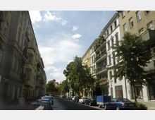 Straßenszene in einem Wohngebiet mit mehrstöckigen, verputzten Altbauhäusern mit Balkonen auf beiden Seiten. Die Straße ist von Bäumen gesäumt, links und rechts parken Autos, und der Himmel ist teilweise bewölkt.