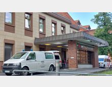 Eingangsbereich einer Notaufnahme mit einem Vordach aus Metall und Backsteinwänden, daneben geparkte Krankenwagen und ein weißer Kleinbus. Das Gebäude hat sandfarbene Fassaden mit rotbraunen Fenstereinfassungen und ein Dach mit roten Ziegeln.