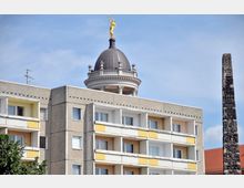 Wohngebäude mit grauen und gelb-weißen Fassadenelementen im Vordergrund, darüber eine Kuppel mit Balustrade und goldener Statue, im Hintergrund ein verzierter Obelisk und ein Dach mit roten Ziegeln.