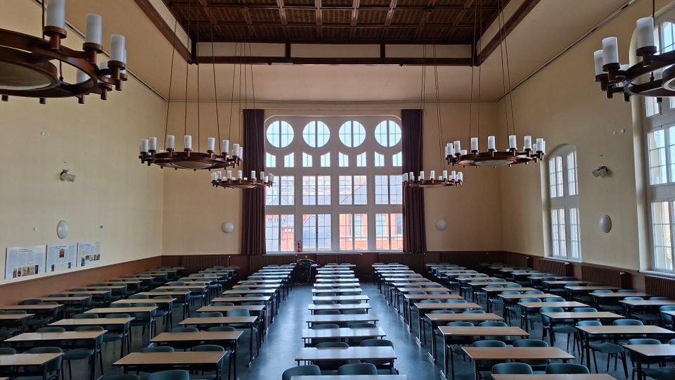 Großer Hörsaal mit hohen Decken, mehreren runden Kronleuchtern und langen Reihen aus Tischen und Stühlen. An der Stirnseite befindet sich ein großes Fenster mit runden Oberlichtern, seitlich hohe Fenster und an den Wänden Informationstafeln.
