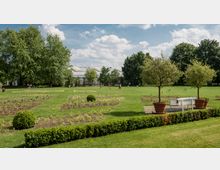 Weitläufige Gartenlandschaft mit gepflegtem Rasen, bepflanzten Blumenbeeten und einer weißen Sitzbank zwischen zwei großen Pflanzenkübeln. Im Hintergrund sind hohe Bäume und ein Gebäude mit gewölbten Glasdächern zu sehen.