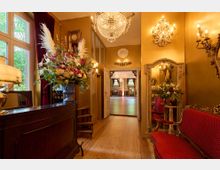Eleganter Eingangsbereich mit goldfarbenen Wänden, verziert durch Kristalllüster, dekorative Wandleuchten und einen großen Spiegel in einem prunkvollen Rahmen. Im Vordergrund stehen ein Tisch mit opulentem Blumenarrangement und rote, gepolsterte Sitzmöbel; im Hintergrund ist ein Saal mit Holzfußboden und Kronleuchtern sichtbar.