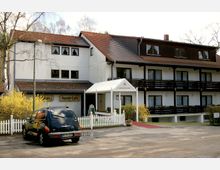 Mehrstöckiges Gebäude eines Hotels mit weißer Fassade, dunklen Holzverkleidungen und roten Ziegeldächern. Vor dem Eingang steht ein schwarzes Auto, neben einem weißen Gartenzaun; blühende Büsche sind im Vordergrund zu sehen.