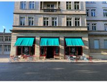 Fassade eines mehrstöckigen Gebäudes mit großen Fenstern und drei Türkisen Markisen im Erdgeschoss, darunter stehen bunte Tische und Stühle. Der Bereich wirkt wie ein Café im Außenbereich, umgeben von einer offenen gepflasterten Fläche.