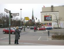 Straßenkreuzung mit Verkehr in einer städtischen Umgebung. Im Hintergrund ist eine Kirche mit einem markanten spitzen Turm sichtbar, daneben ein Gebäude mit großen Werbeplakaten an der Fassade.