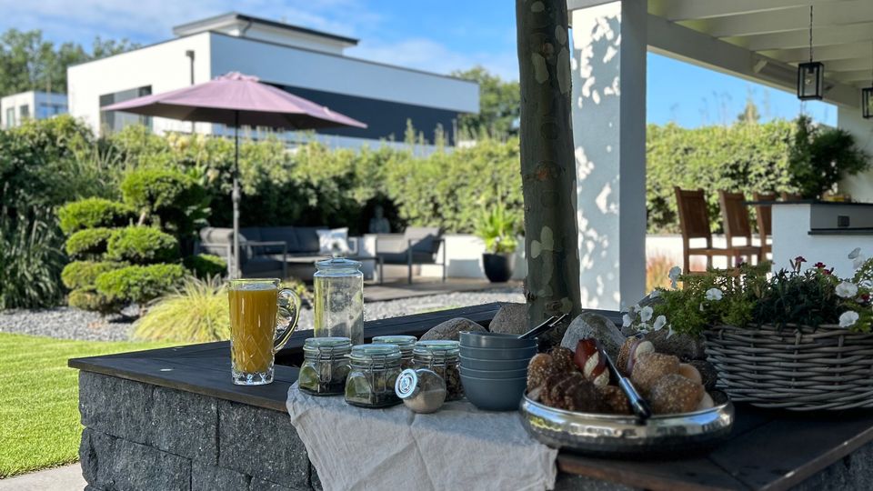 Überdachte Terrasse in einem modernen Garten mit einer Sitzlounge, Sonnenschirm und gepflegten Hecken im Hintergrund. Im Vordergrund steht ein gemauerter Außenbereich mit Ablagefläche unter einem Baum, darauf Gläser und Tabletts mit Frühstücks- oder Snackutensilien.