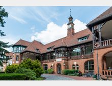 Backsteingebäude im historischen Stil mit roten Ziegeldächern, Fachwerkelementen und einem kleinen Turm mit Uhr. Vor dem Gebäude befinden sich Bäume, Sträucher, ein gepflasterter Weg und mehrere Fahrräder.