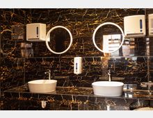 Waschraum mit zwei weißen ovalen Waschbecken auf einer schwarz-goldenen Marmorarbeitsfläche, zwei runden Spiegeln mit Beleuchtung an der Wand und Spendern für Seife und Papierhandtücher. Die Wände sind mit schwarzem Marmor mit goldenen Adern verkleidet.