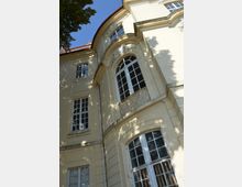 Gelbes historisches Gebäude mit großen, weißen Sprossenfenstern und barocken Dekorationen, darunter geschwungene Fassadenelemente und ein kunstvoll verziertes schmiedeeisernes Geländer. Im Vordergrund wirft ein Baum Schatten auf die Fassade, und der Himmel ist blau.