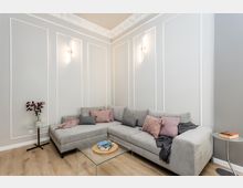 Modernes Wohnzimmer mit hellgrauer Eckcouch, Glas-Couchtisch und kleinen Beistelltischen auf Holzfußboden. Hohe Wände mit weißen Zierleisten und Stuckleiste an der Decke sowie zwei schmale Wandlampen prägen den Raum.