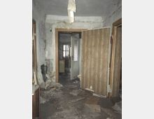 Ein verlassener, heruntergekommener Innenraum mit abblätternder Tapete, kaputter Decke und Müll auf dem Boden. Eine teilweise geöffnete Holztür und eine Glastür im Hintergrund führen zu weiteren Räumen.