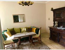 Wohnzimmer mit klassischem Interieur: Ein grünes Sofa mit farbigen Kissen und ein Holztisch mit Tischläufer und Kerzenständer stehen auf einem flauschigen Teppich. Rechts steht eine massive, verzierte Holzanrichte mit Gläsern, darüber hängt ein dekorativer Kronleuchter.