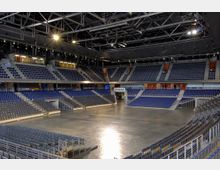 Innenansicht einer großen Mehrzweckhalle mit leeren Sitzreihen in mehreren Ebenen, einer zentralen freien Fläche mit Betonboden und einer sichtbaren Stahlkonstruktion an der Decke. Der Raum ist hell beleuchtet, und es gibt Logen und Tribünenplätze.
